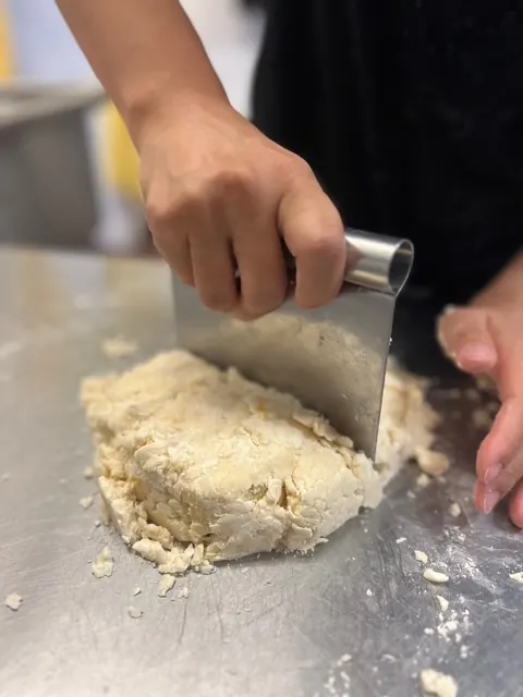 Hands lifting a bench scraper through a mound of fresh dough on a steel table.