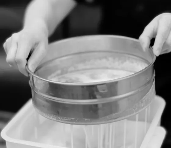 Gloved hands sifting flour into a tub of batter in the shop kitchen.