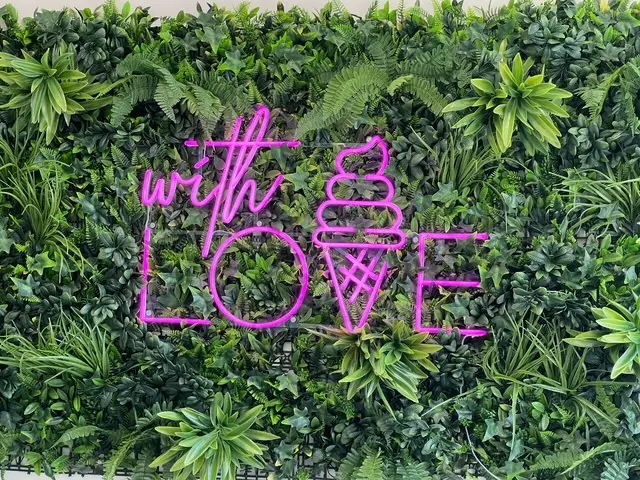 The shop's magenta neon sign reading with LOVE. mounted on a wall of green foliage.